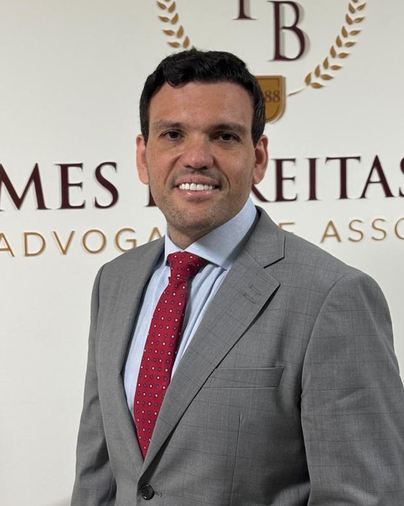Fábio Gomes de Freitas Bastos, de terno cinza e gravata vermelha, em frente ao logotipo do escritório